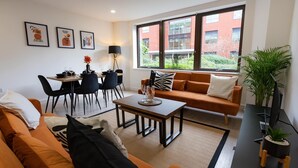 Un salon moderne avec un canapé, une table basse et un coin repas. On y trouve des œuvres d’art encadrées au mur, une grande fenêtre donnant sur des immeubles et une plante en pot.