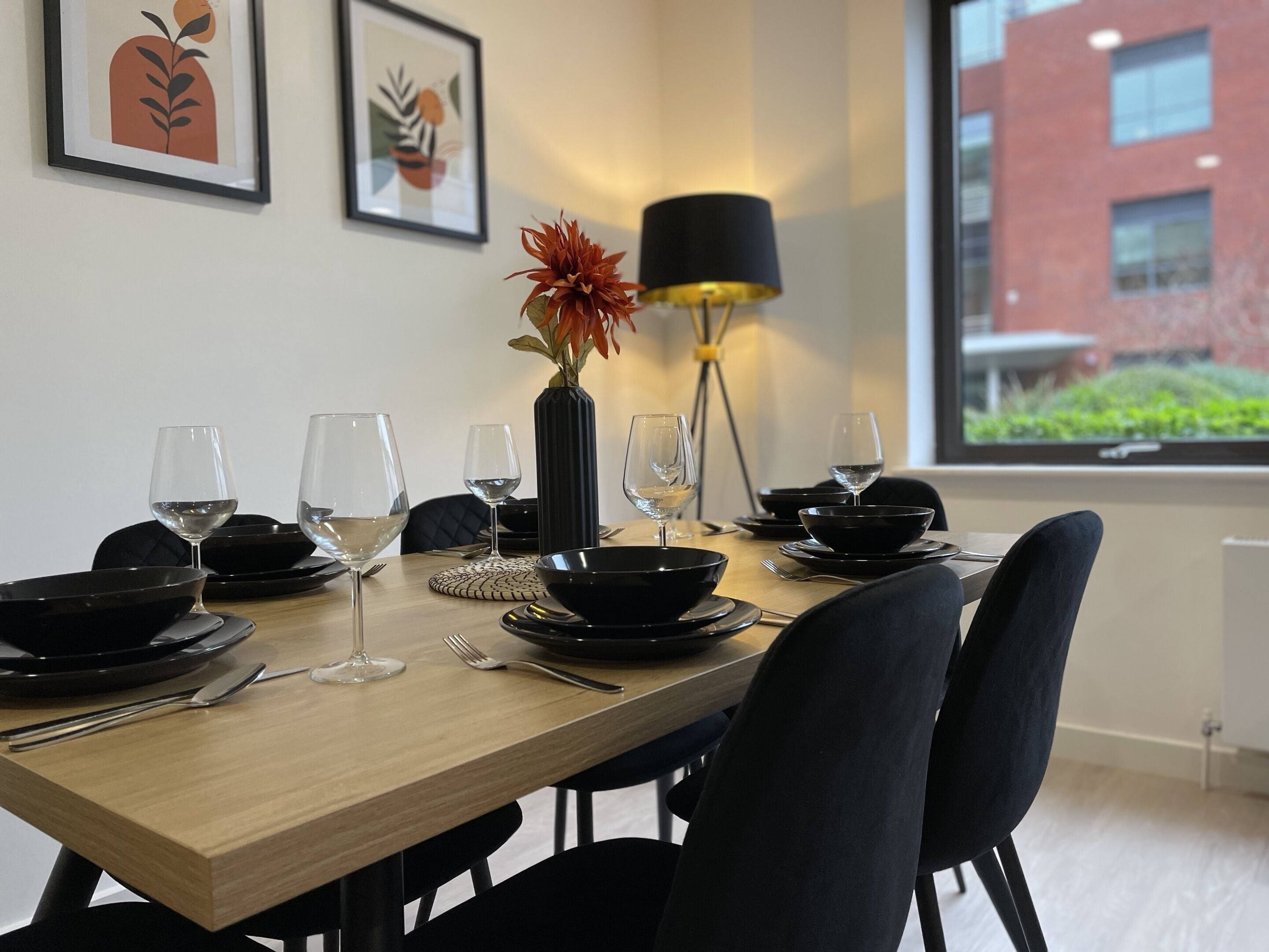 A dining table set with black dinnerware, wine glasses, and a vase with a flower.