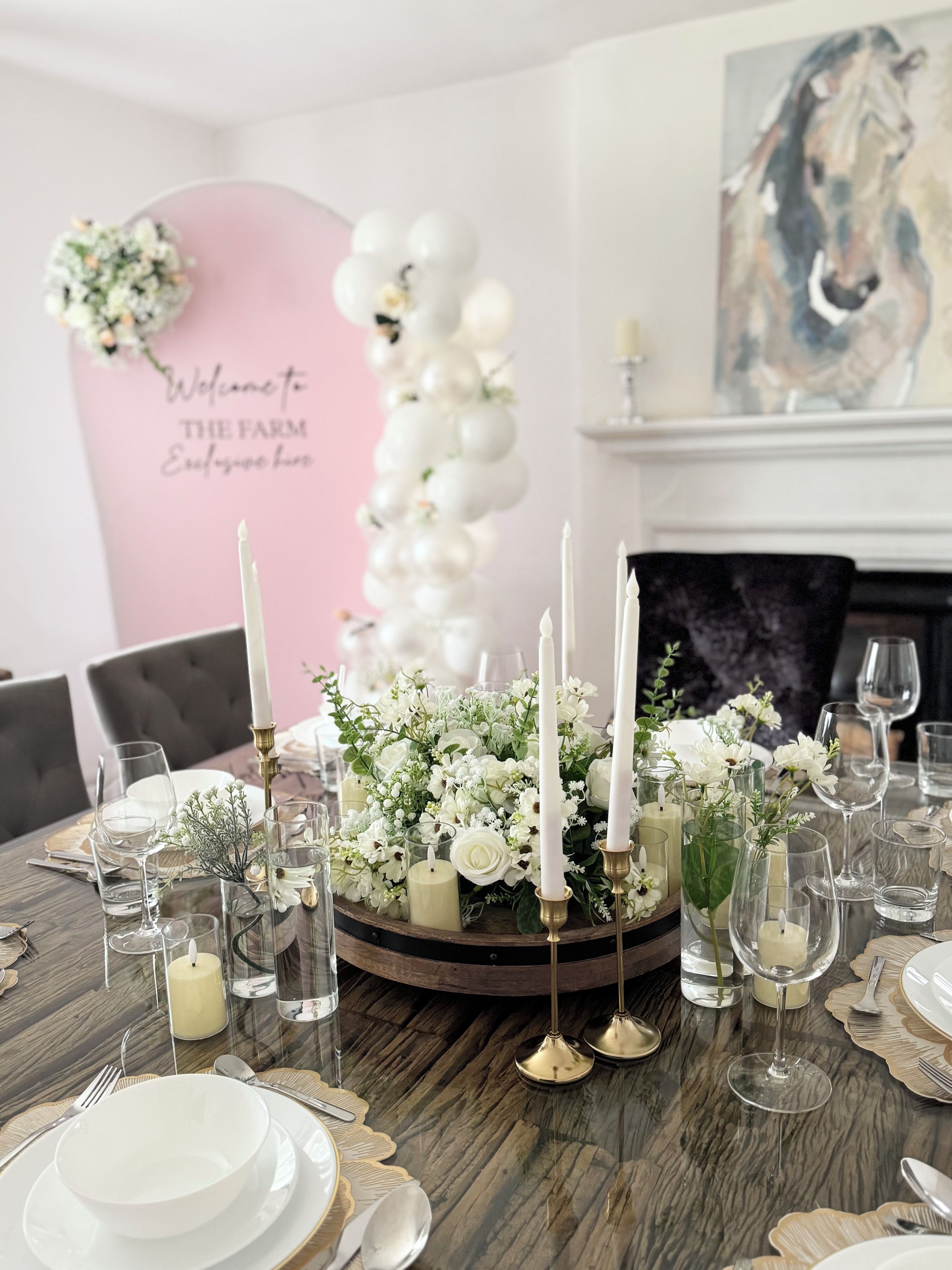 A dining table set with white plates, glasses, and candles, surrounded by floral arrangements and a decorative wall with a welcome sign.