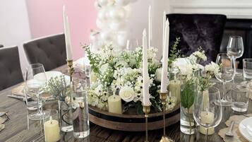 A dining table set with white plates, glasses, and candles, surrounded by floral arrangements and a decorative wall with a welcome sign.