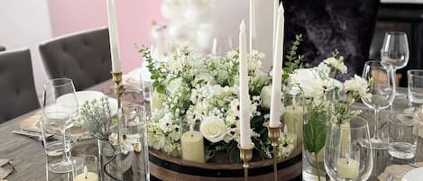 A dining table set with white plates, glasses, and candles, surrounded by floral arrangements and a decorative wall with a welcome sign.