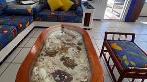 Ein Wohnzimmer mit großem Fenster zum Meer, einem farbenfrohen Sofa, einem kleinen Tisch und einem dekorativen Aquarium.