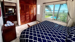 Ein Schlafzimmer mit Bett, Deckenventilator, Fenster mit Meerblick, Holzschrank und Badezimmer mit Spiegel und Handtuchhalter.