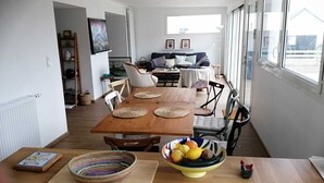 A spacious living area with a wooden dining table, chairs, and a sofa. There is a large window providing natural light and a view of the outdoors.