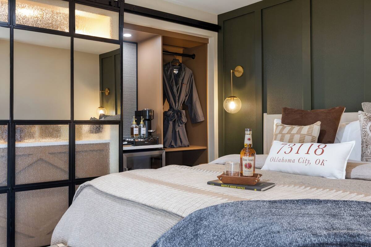 Habitación de hotel con una cama, una mesita de noche con una botella y un libro, una bata colgada en un perchero y una almohada con el nombre de una ciudad.