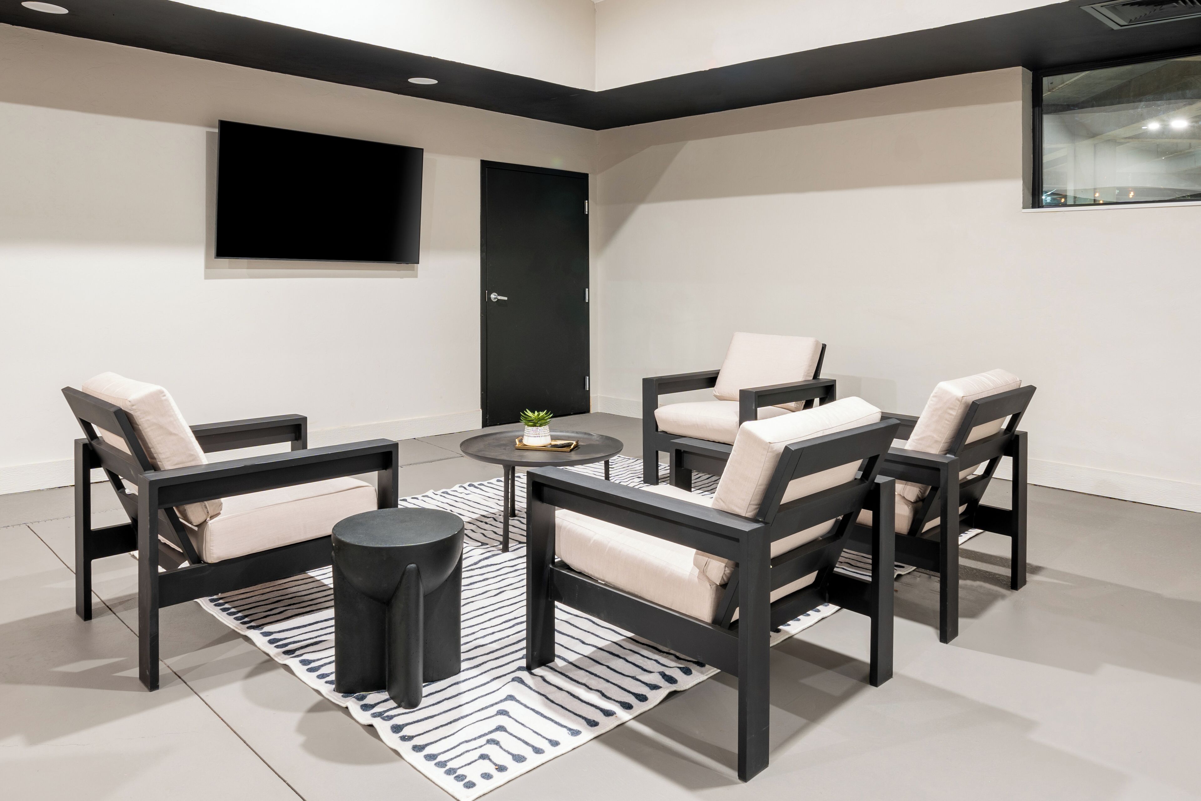 Una sala de estar moderna con un sofá negro, una mesa de centro redonda y un sillón decorativo negro. Se ve una puerta negra y una ventana al fondo.