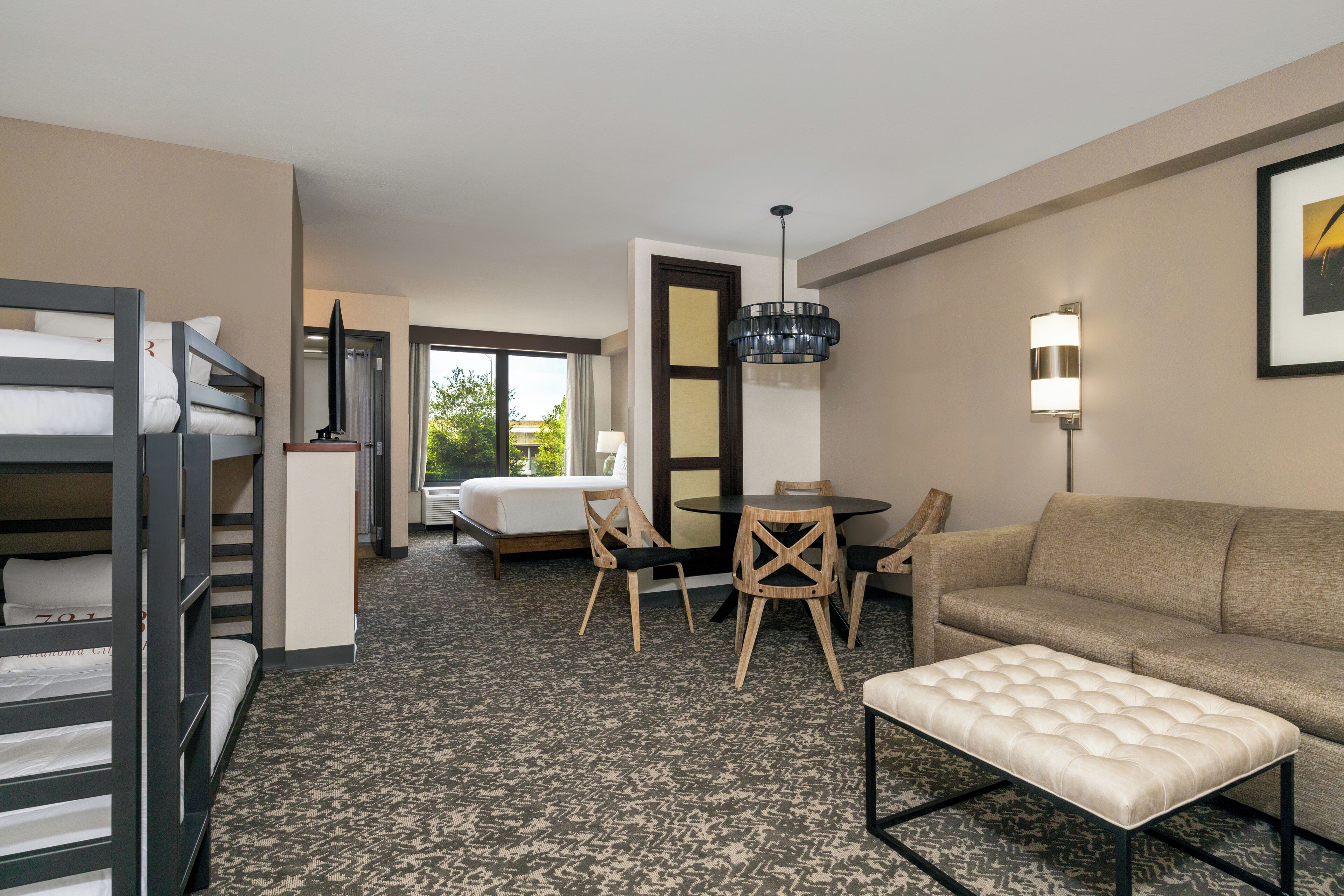 A hotel room with bunk beds, a sofa, a dining table with chairs, a chandelier, and a large window.