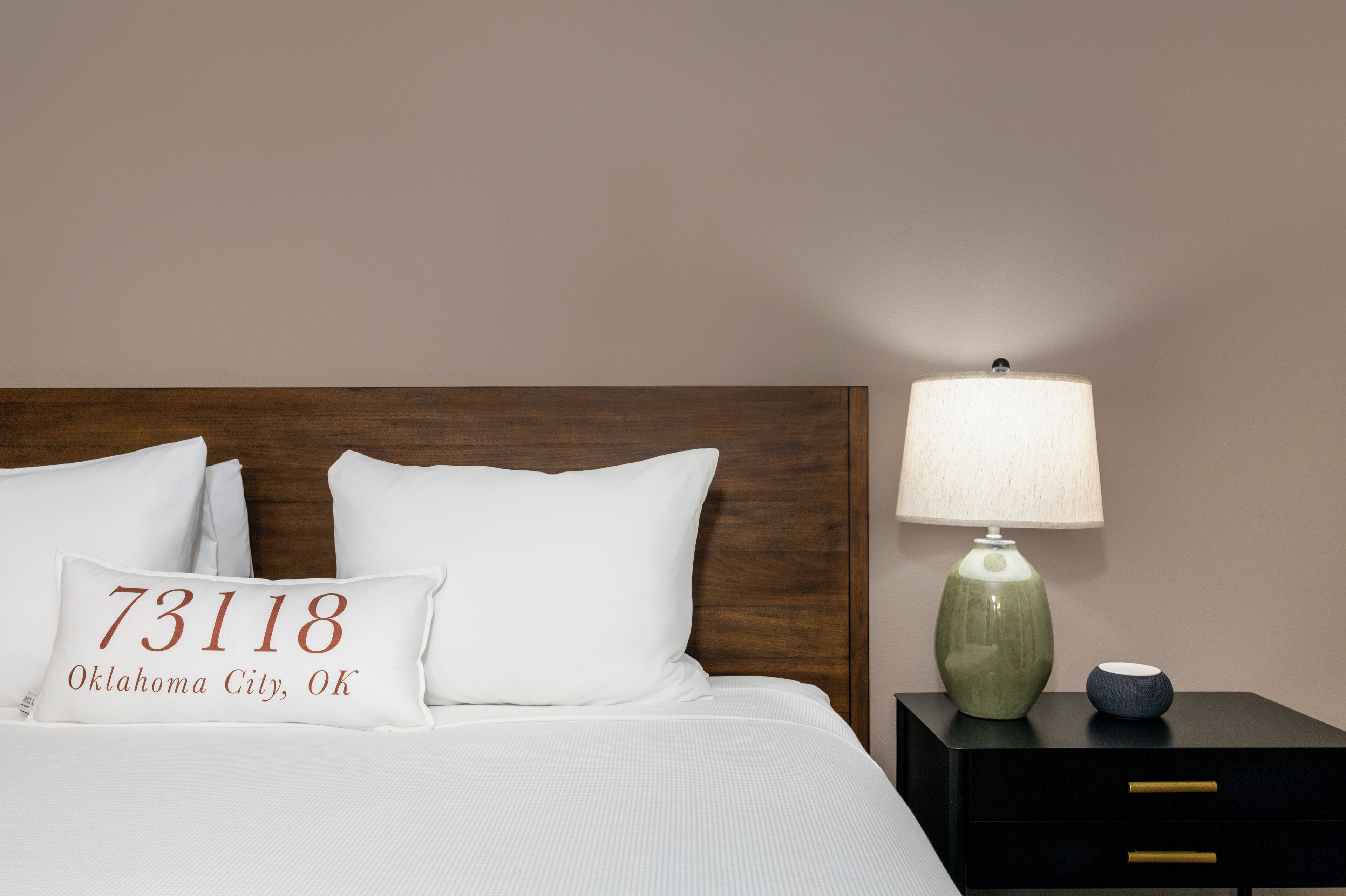 Ein ordentlich bezogenes Bett mit weißer Bettwäsche und einem Kissen mit der Aufschrift '73181 Oklahoma City, OK'. Eine grüne Keramiklampe und eine kleine, blaue Schale stehen auf einem schwarzen Nachttisch neben dem Bett.