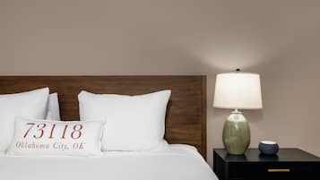 Una cama impecablemente tendida con ropa blanca y una almohada que muestra '73118 Oklahoma City, OK'. Una lámpara de cerámica verde y un pequeño cuenco azul están sobre una mesita de noche negra junto a la cama.
