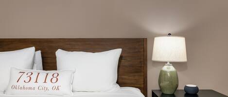 A neatly made bed with white bedding and a pillow displaying '73118 Oklahoma City, OK'. A green ceramic lamp and a small blue bowl are on a black nightstand beside the bed.
