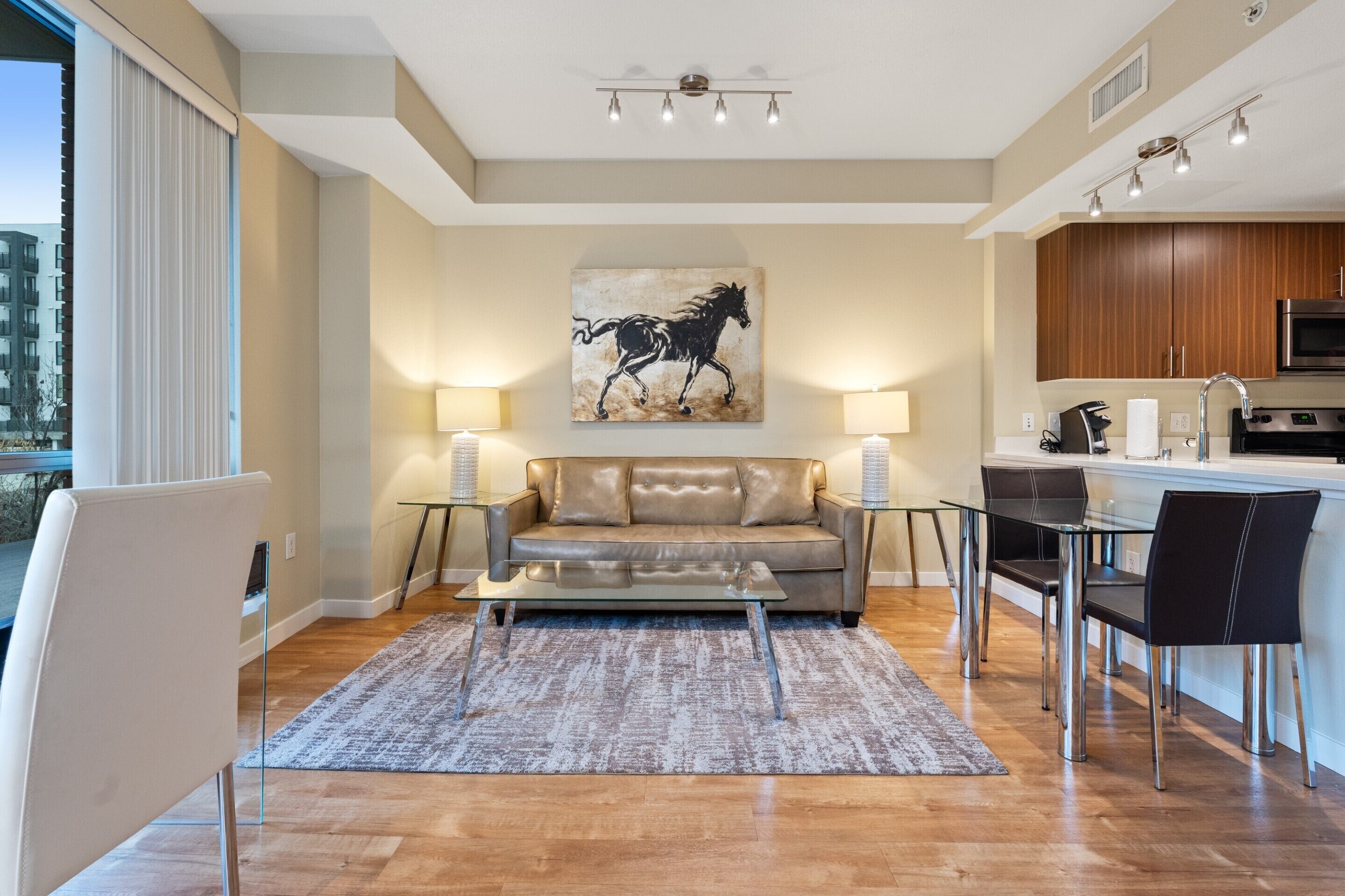 A modern living room with a sofa, coffee table, and dining area. There is a kitchen with a microwave and cabinets. A painting of a horse is on the wall.