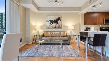 A modern living room with a sofa, coffee table, and dining area. There is a kitchen with a microwave and cabinets. A painting of a horse is on the wall.