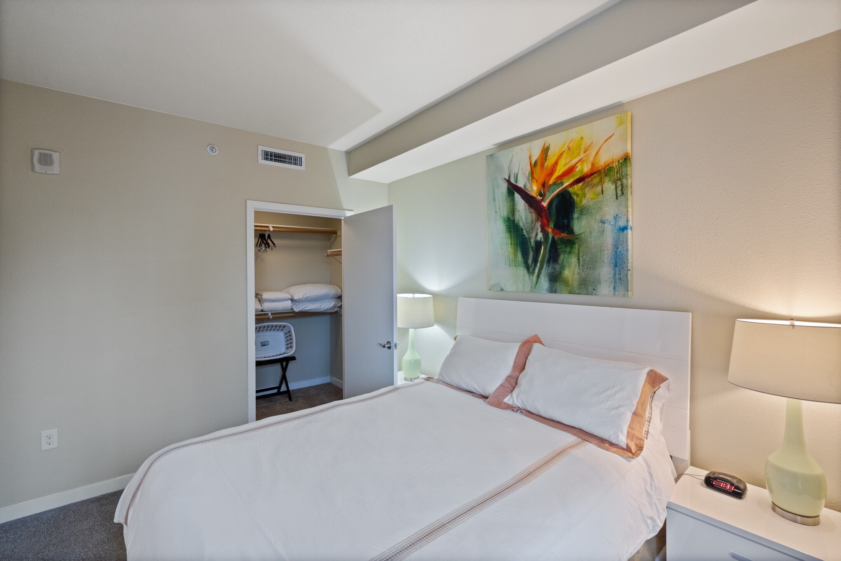 A hotel room with a large bed, a nightstand with a lamp, a painting on the wall, and a closet with hanging clothes.