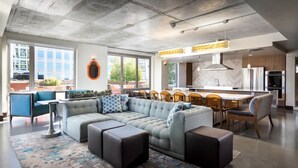 Un salon moderne avec un canapé d’angle, un coin repas avec des chaises, une cuisine dotée d’un grand îlot central et une vue sur la ville grâce à de grandes fenêtres.