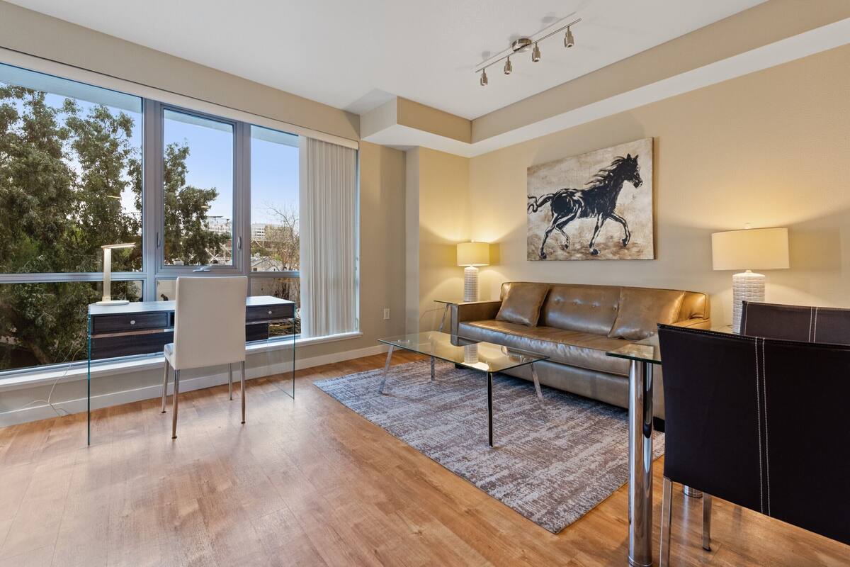 Un salon moderne avec une table basse en verre, un canapé en cuir et une grande fenêtre donnant sur des arbres et des immeubles.