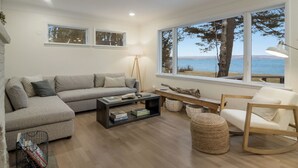 A modern living room with a large window offering a view of the sea, a grey sectional sofa, a wooden rocking chair, and a coffee table with books. - Wander Cottage | Lakefront | Outdoor Shower | Deck (Maple City)