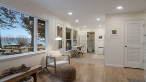 A spacious room with a large window, a wooden bench, a white chair, and a small ottoman. - Wander Cottage | Lakefront | Outdoor Shower | Deck (Maple City)
