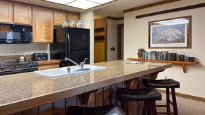 A kitchen with a granite countertop, a black refrigerator, wooden cabinets, a microwave, a stove, and a sink. There are two stools and a shelf with books and decorative items. - Mammoth Getaway, Short walk to the Village and steps to Canyon Lodge shuttle. (Mammoth Lakes)