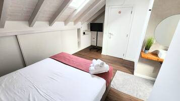 A bedroom with a bed, a red blanket, a white towel, a small rug, a wooden floor, a white door, a black TV stand, and a mirror.