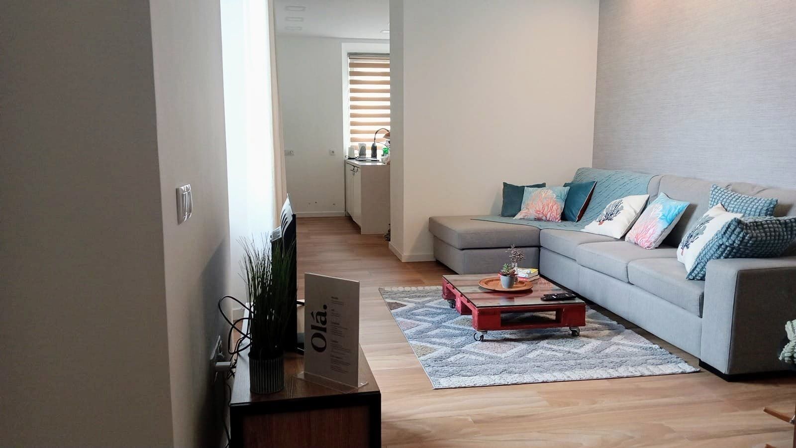 Ein Wohnzimmer mit grauem Sofa, einem Couchtisch und einem Teppich. Auf dem Couchtisch liegt ein Schild und links steht eine Pflanze.