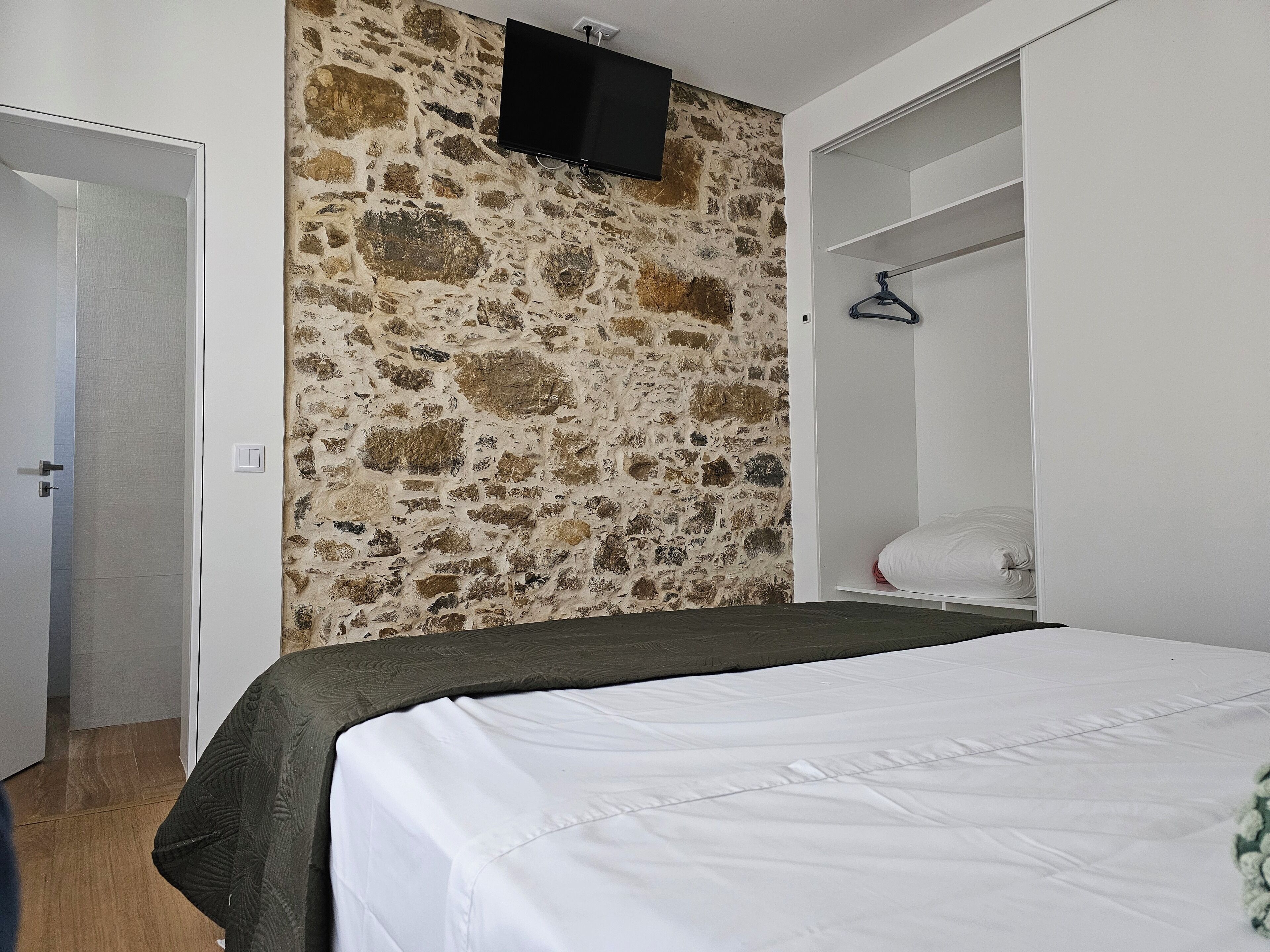 Ein Schlafzimmer mit Stehwand, einem Bett mit weißer Bettwäsche, einem Kleiderschrank und einem an der Wand montierten Fernseher.