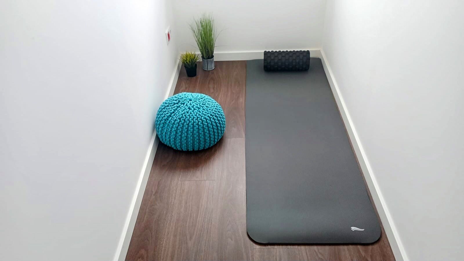 A yoga mat and a blue knitted ottoman in a corner of a room with wooden flooring and white walls.