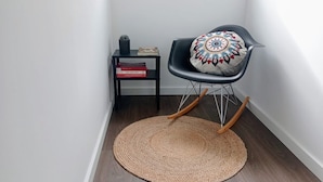 A corner room with a rocking chair, a woven rug, a small black side table with books, and a decorative pillow. - SurfSoul B&B (Peniche)