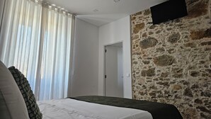A bedroom with a bed, a window with curtains, a stone wall, and a mounted TV. - SurfSoul B&B (Peniche)