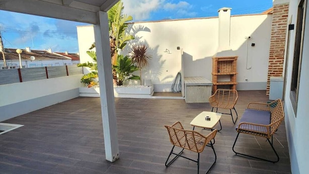 A covered patio with dark wooden flooring, wicker furniture, and a white wall with a chimney. - SurfSoul B&B (Peniche)