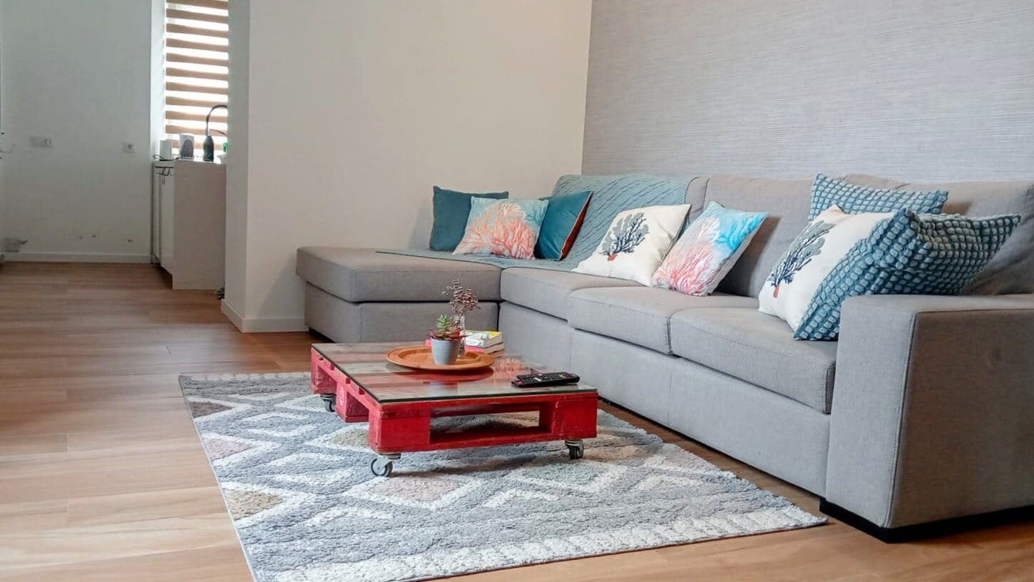 A modern living room with a grey sectional sofa, colorful cushions, a red coffee table with a tray, and a patterned rug.