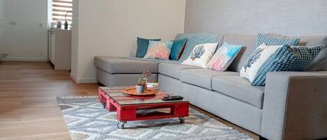 A modern living room with a grey sectional sofa, colorful cushions, a red coffee table with a tray, and a patterned rug.