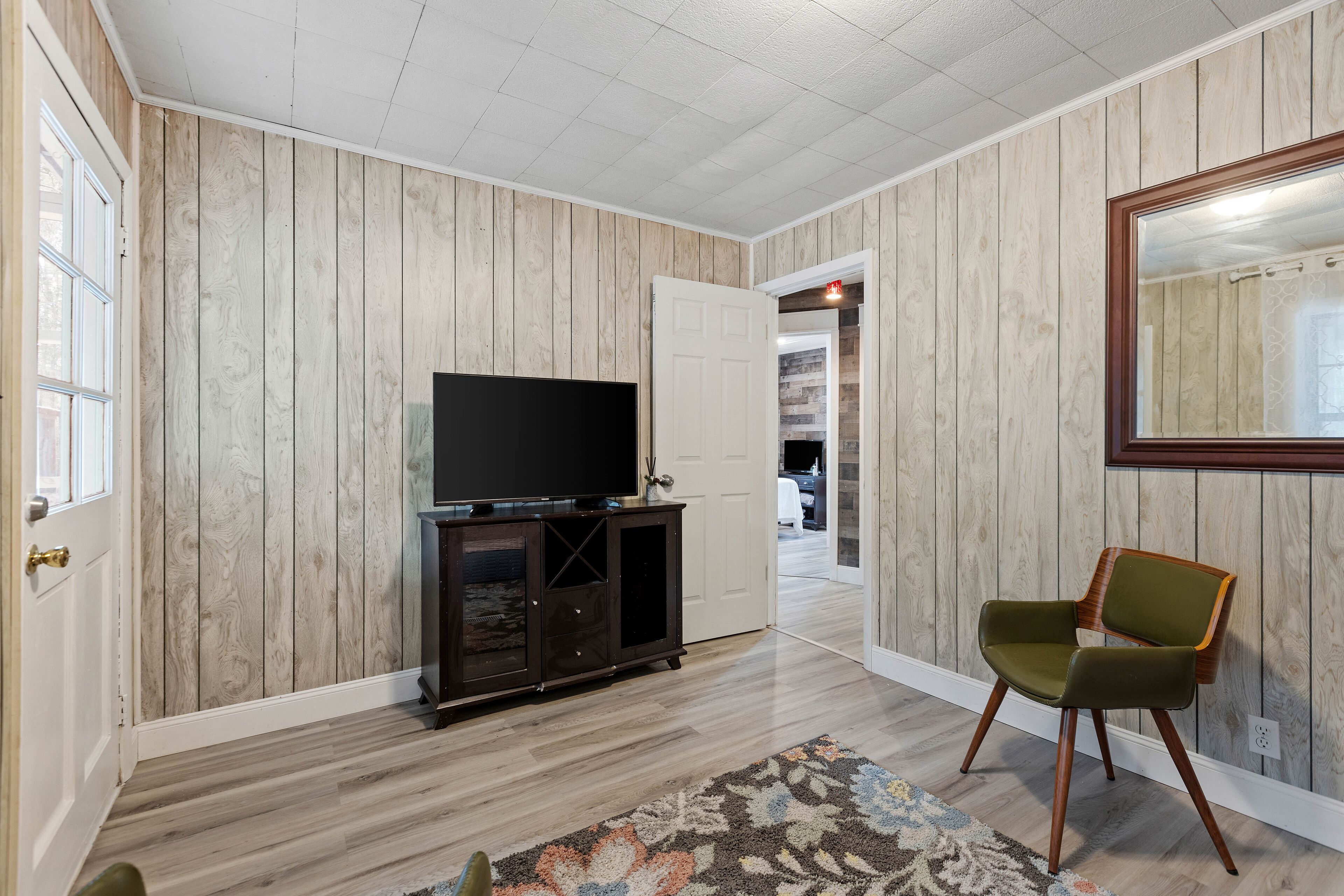 A room with a wooden paneled wall, a television on a cabinet, a green chair, and a patterned rug.