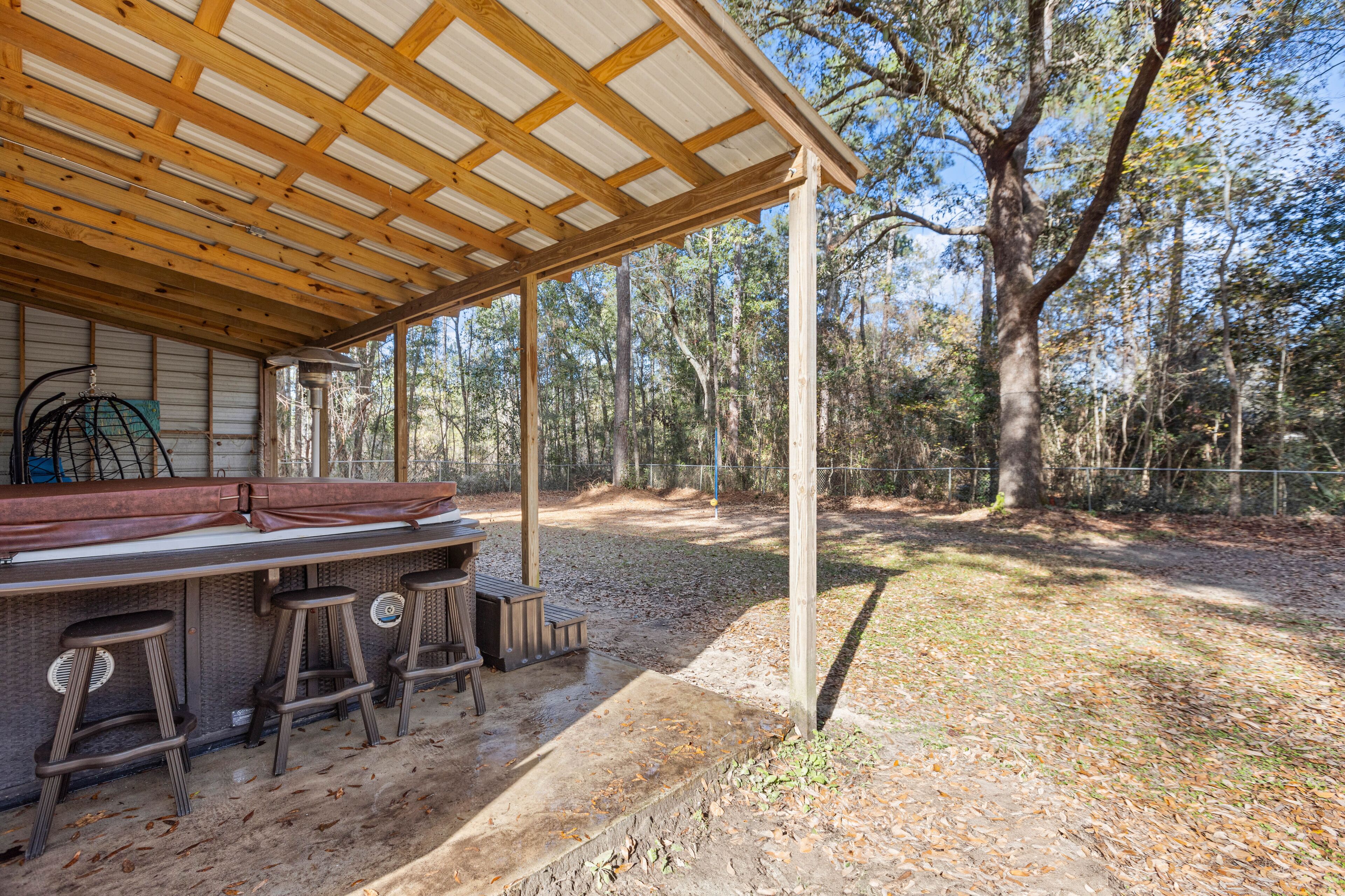 Un espace bar extérieur abrité, doté de poutres en bois et d’un sol en béton, entouré d’arbres et d’une cour clôturée.