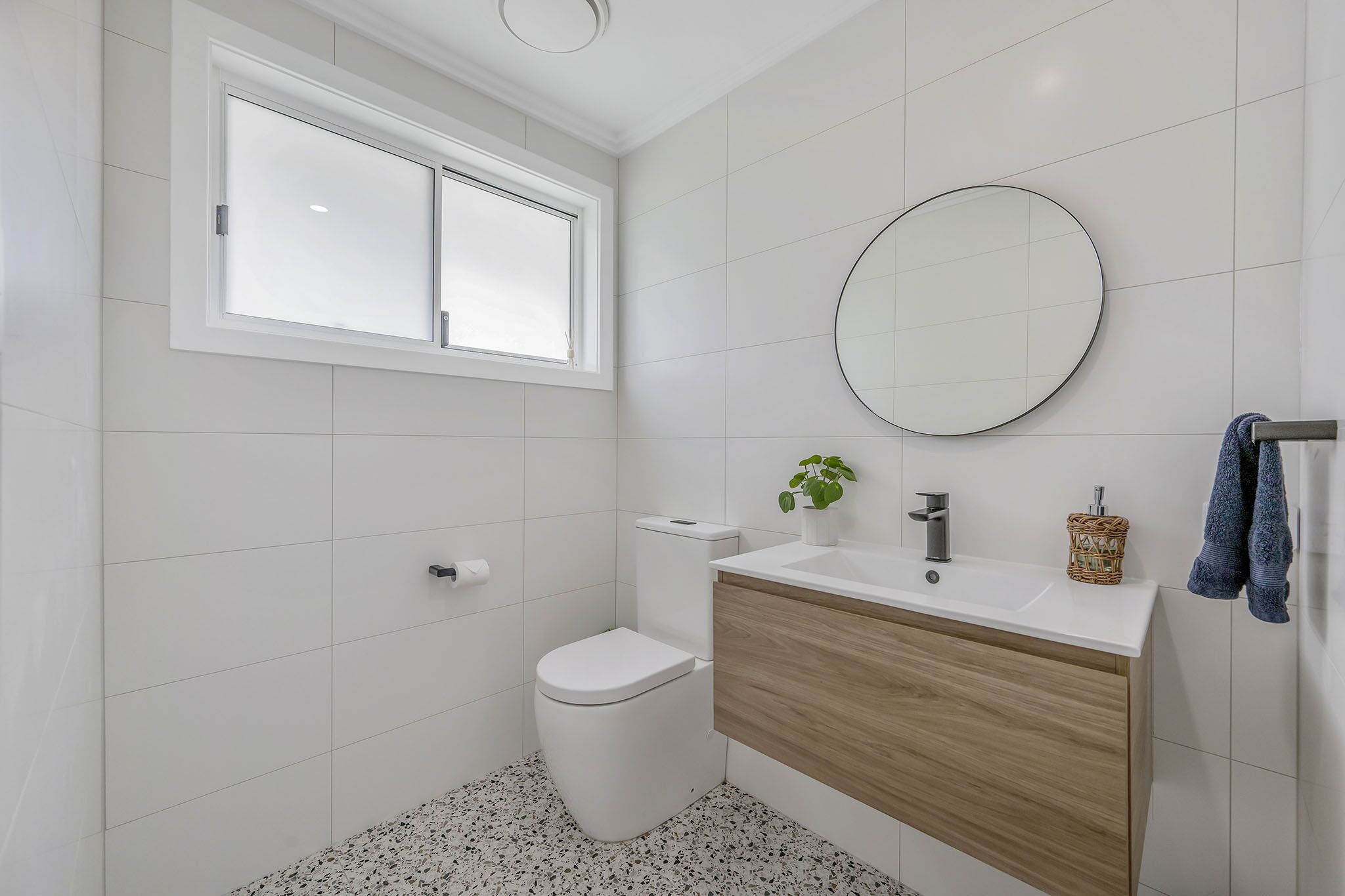 Une salle de bain moderne avec des carreaux blancs, des toilettes, un lavabo avec un meuble-vasque en bois, un miroir rond et une fenêtre.