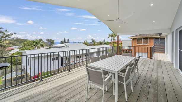 Une terrasse en bois avec une table et des chaises, un ventilateur de plafond et une vue sur l’océan et les maisons voisines.