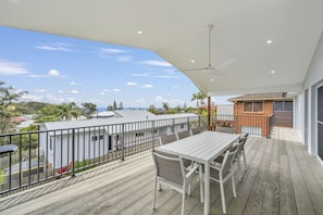 Une terrasse en bois avec une table et des chaises, un ventilateur de plafond et une vue sur l’océan et les maisons voisines.