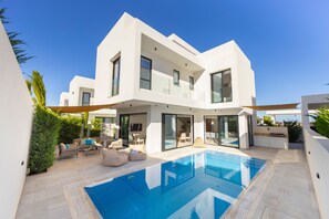Et moderne hus med to etager, en swimmingpool, udendørs møbler og en stor glasdør.