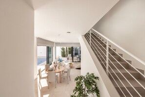 A modern interior with a staircase, a dining area with a table and chairs, and a view through large windows.