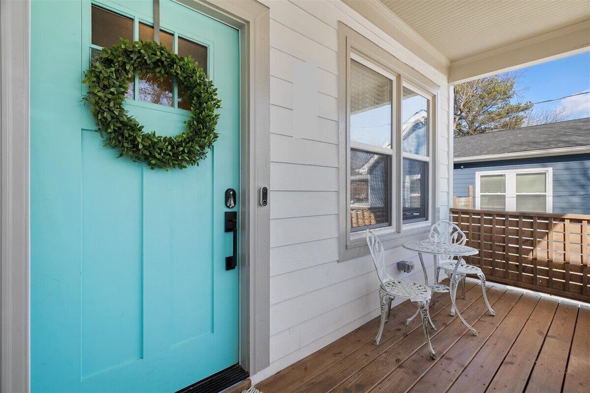 A turquoise door with a wreath, a white porch with two white metal chairs, and a wooden deck.
