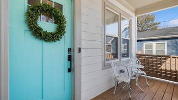 A turquoise door with a wreath, a white porch with two white metal chairs, and a wooden deck.