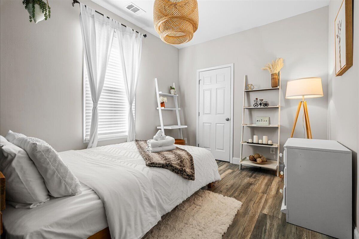 Une chambre avec un lit, des rideaux blancs, une porte blanche, une étagère blanche ornée d’objets décoratifs, une lampe blanche et un luminaire suspendu en osier.