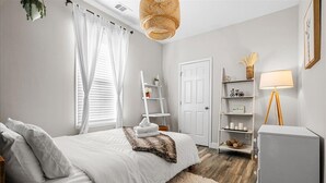 Ein Schlafzimmer mit einem Bett, weißen Vorhängen, einer weißen Tür, einem weißen Regal mit Dekoration und einer weißen Lampe sowie einem hängenden Weidenlampenschirm.