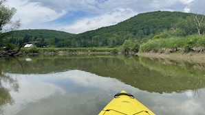 A calm river with a yellow kayak in the foreground, surrounded by lush green hills and a partly cloudy sky.