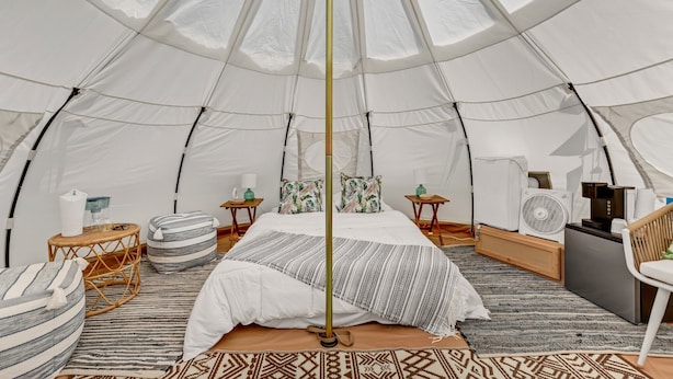 A spacious tent with a bed, pillows, a fan, a small table, and a chair.