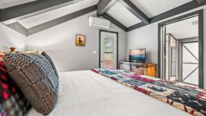 Ein Schlafzimmer mit schrägen Wänden, einem Bett mit Quilzdecke, einem Fernseher auf einem Standfuß und einer Tür zu einem Außenbereich.