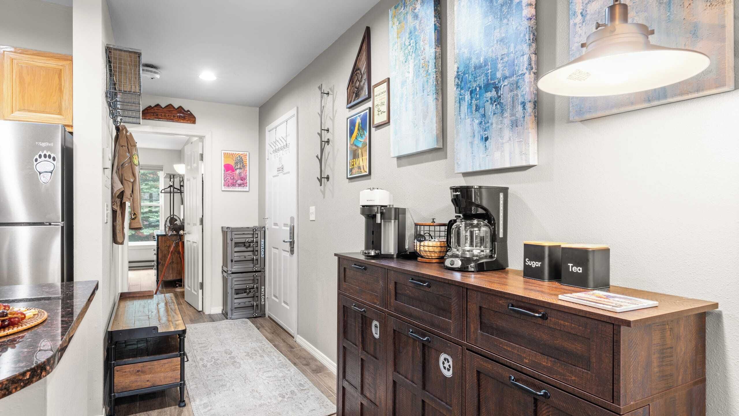 A modern kitchen with a refrigerator, coffee maker, and cabinets. There are artwork on the walls and a rug on the floor.