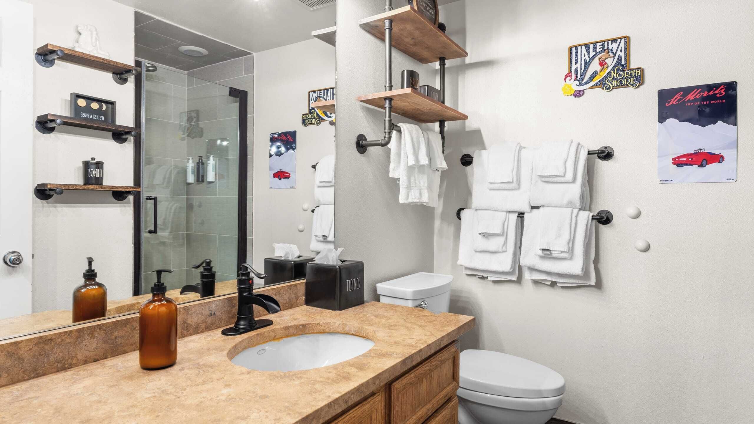 Une salle de bain moderne avec un grand miroir, un lavabo doté d’un plan de toilette en marbre, des toilettes et un espace douche. Des serviettes sont accrochées au mur et divers produits de toilette sont disposés sur des étagères.