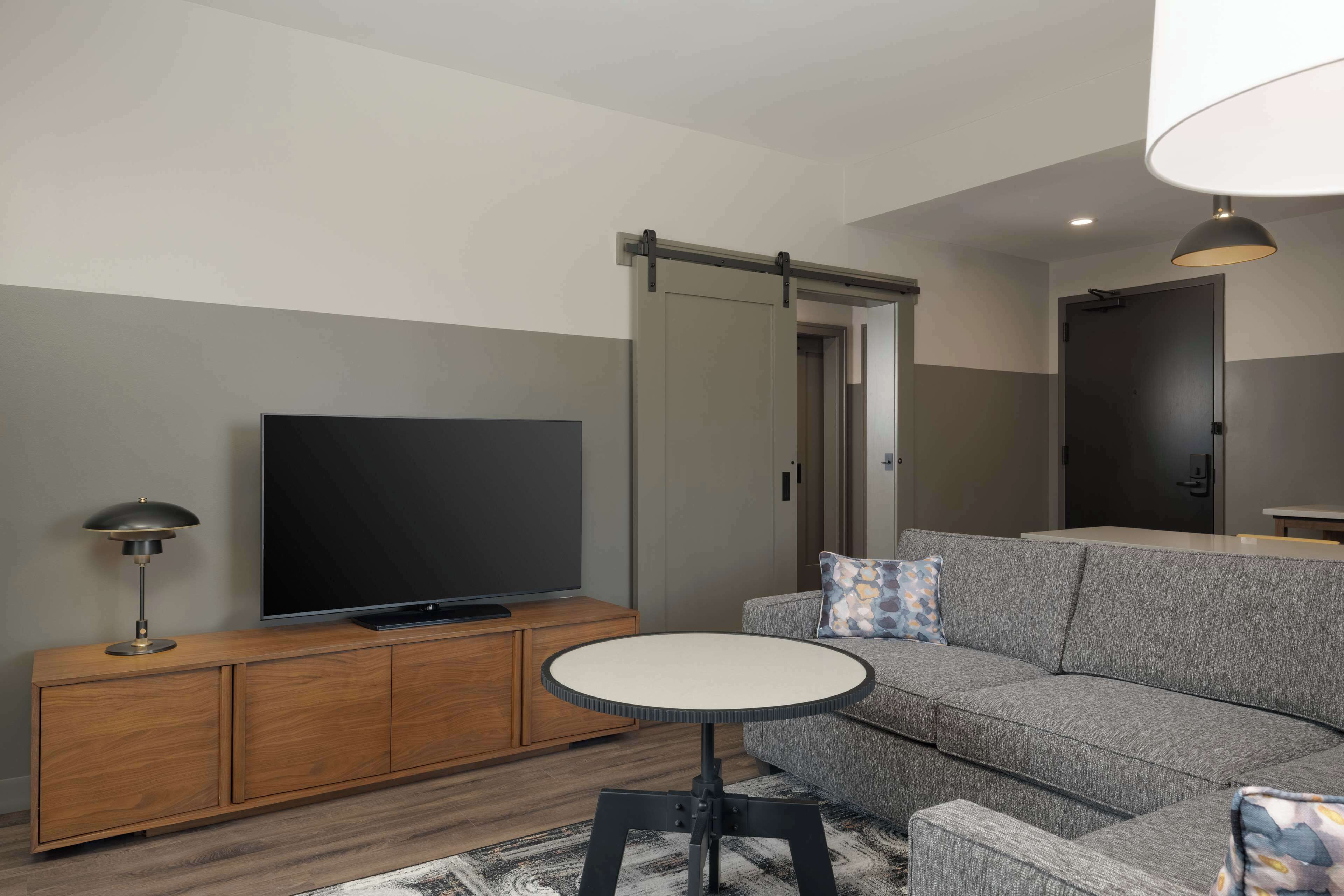 A modern living room with a grey sofa, a flat-screen TV on a wooden cabinet, a round table, and a sliding door closet.