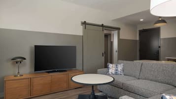 Uma sala de estar moderna com um sofá cinzento, uma televisão de tela plana sobre um móvel de madeira, uma mesa redonda e um guarda-roupa com portas deslizantes.