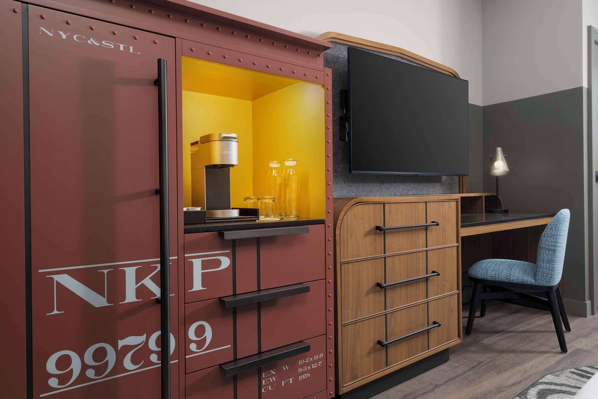 A compact hotel room with a kitchenette, a flat-screen TV, a desk, and a chair.
